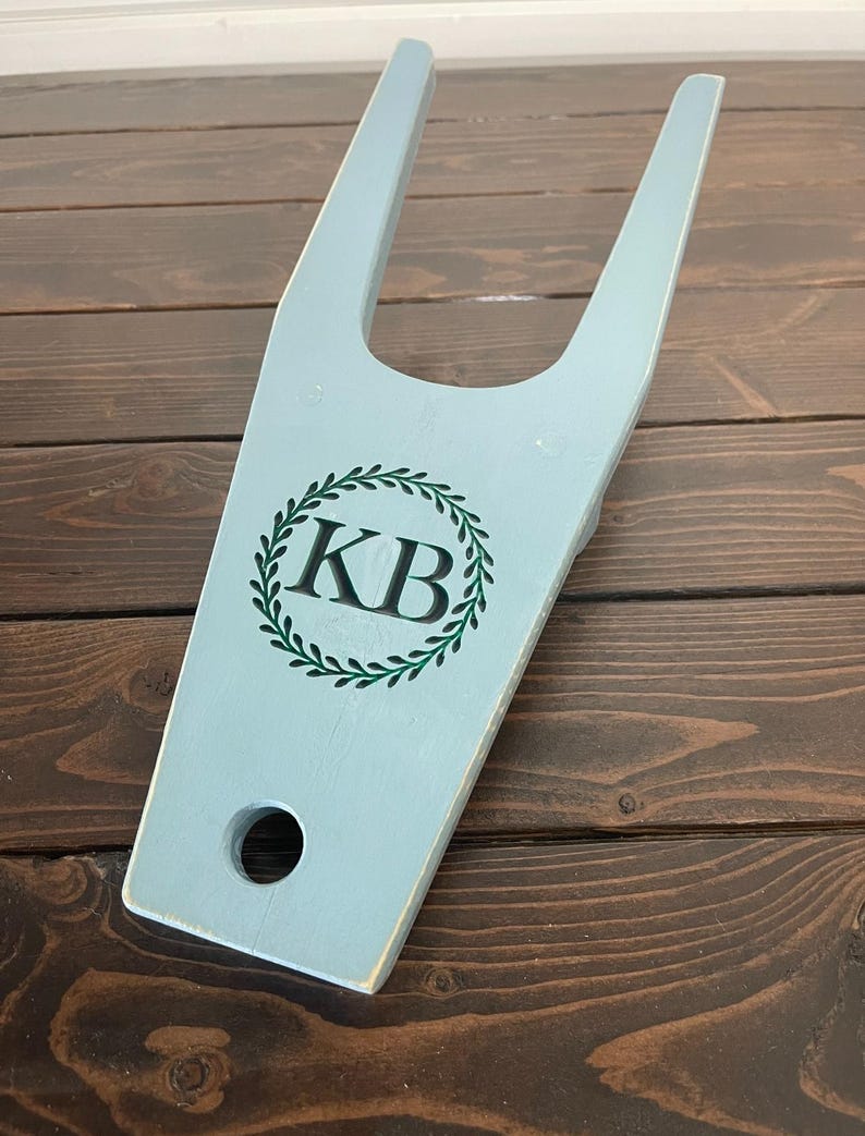 May include: A light blue wooden boot jack with a green wreath and the initials "KB" in the center.