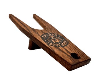 May include: A wooden boot jack with a brown stain finish. The boot jack has a cutout for the heel of a boot and a handle with a rounded end. The boot jack is engraved with the words "Fat Dog Boot Jacks" and a bulldog logo.