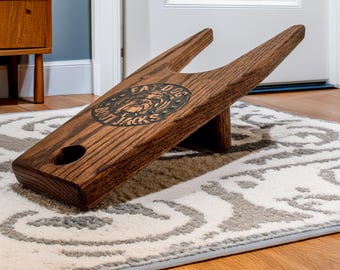 May include: A wooden boot jack with a dark brown stain. The boot jack has a carved design of a dog's head with the text "Fat Dog Boot Jacks".