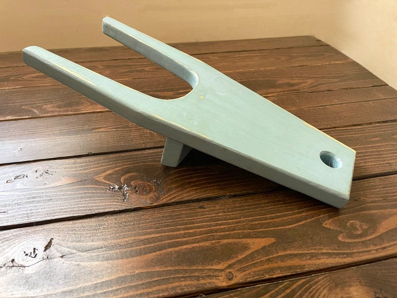 May include: A light blue wooden boot jack with a rounded handle and a hole in the base. The boot jack is sitting on a dark brown wooden surface.