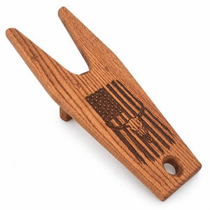 May include: A brown wooden boot jack featuring a laser-engraved design of an American flag combined with a steer skull. The tool has a V-shaped opening at the top for removing footwear and a circular hole at the bottom for hanging storage.