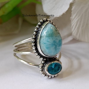 Op de afbeelding: Een zilveren ring met twee blauwe edelstenen. De grotere steen is een traanvormige blauwe kant-agate, en de kleinere steen is een ronde blauwe topaas. De ring heeft een parelmoerontwerp rond de stenen.