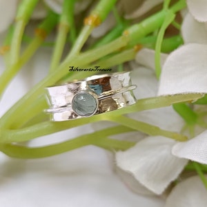 Op de afbeelding: Een draaibare zilveren ring met een lichtblauwe edelsteen. De ring is getextureerd en heeft een gehamerde afwerking. De ring ligt op een witte achtergrond.