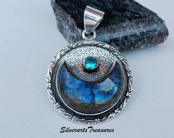 May include: A silver pendant with a crescent moon design featuring a blue labradorite stone and a smaller blue gemstone at the top. The pendant is surrounded by a silver border with a textured design.