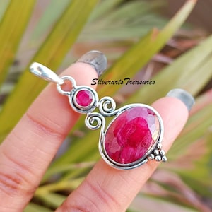 May include: A silver pendant with a large oval ruby gemstone and a smaller ruby gemstone. The pendant has a delicate, swirling design and is set on a silver chain.
