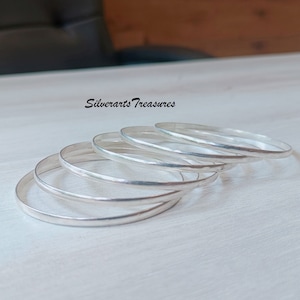 Puede incluir: Cinco pulseras de plata apiladas una encima de la otra sobre una superficie blanca. Las pulseras son todas lisas y brillantes.