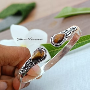 Op de afbeelding: Zilveren armband met twee druppelvormige tijgeroogstenen. De armband is gemaakt van sterling zilver en heeft een delicaat bloemmotief.