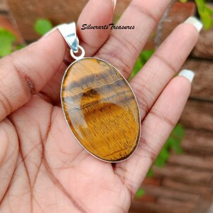 May include: A silver pendant featuring a large oval-shaped tiger's eye gemstone. The gemstone is a rich golden brown color with a shimmering, chatoyant effect.