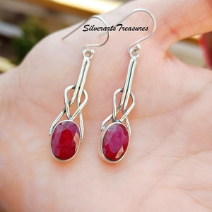 Puede incluir: Un par de pendientes de plata con una piedra preciosa de rubí rojo. Los pendientes presentan un delicado diseño de nudo y cuelgan de un gancho.