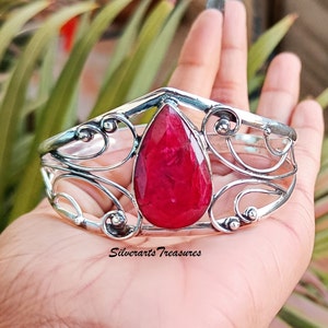 Könnte beinhalten: Ein silbernes Armreif-Armband mit einem großen, tropfenförmigen Rubin-Edelstein, der in der Mitte eingesetzt ist. Das Armband hat ein zartes, verziertes Design mit Spiralen und Kurven.