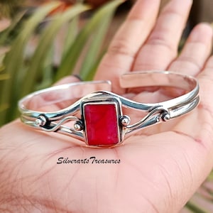 Puede incluir: Un brazalete de plata con una piedra preciosa roja engastada en un marco rectangular. El brazalete tiene un diseño delicado y fluido con dos bandas y pequeños detalles decorativos.