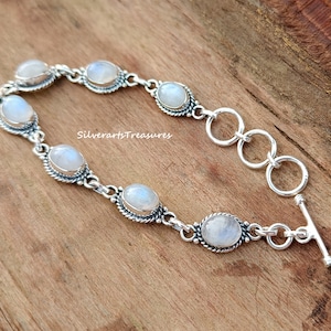 以下が含まれることがあります： トグル留め具が付いたシルバーブレスレットは、シルバーのベゼルにセットされた6つのオーバルムーンストーンジェムストーンが特徴です。ブレスレットは木製の表面にあります。Silverartstreasures.