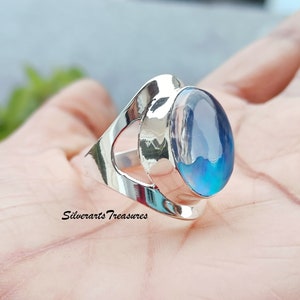 Puede incluir: Un anillo de plata con una gran piedra preciosa ovalada azul engastada en un diseño abierto único. El anillo está en una mano, pero la mano no es visible. El texto "Silverarts Treasures" es visible en el anillo.