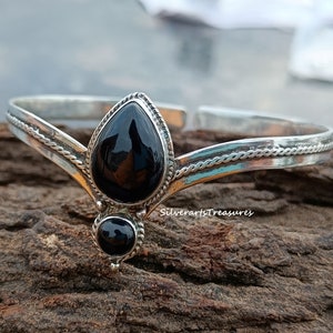 May include: A silver bangle bracelet with two black onyx stones. The larger stone is teardrop shaped and the smaller stone is round. The bracelet has a braided design.