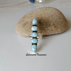 May include: A silver ring with six light blue gemstone stones set in a vertical row. The stones are oval shaped and have a pearlescent finish. The ring is on a light brown rock.