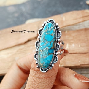 Puede incluir: Un anillo de plata con una gran piedra ovalada de turquesa engastada en un engaste de plata decorativo. La piedra tiene un color azul y verde con vetas marrones que la atraviesan.