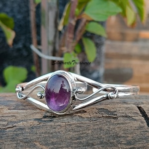 May include: A silver bracelet with a purple amethyst gemstone set in a delicate, ornate design. The bracelet features a curved, open-ended band with a central oval-shaped stone.