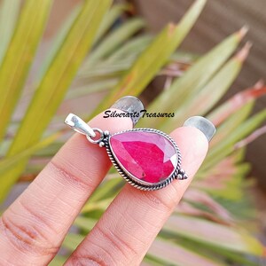 Puede incluir: Un colgante de rubí en forma de lágrima engastado en plata de ley. El rubí es de color rojo intenso y está rodeado por un engaste de plata. El colgante está en una cadena de plata.