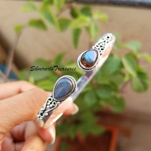 May include: A silver bangle bracelet featuring two teardrop-shaped labradorite gemstones. The bracelet has a delicate, ornate design.