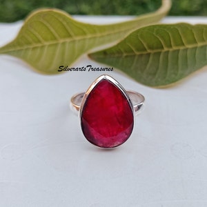 May include: A silver ring featuring a teardrop-shaped ruby gemstone. The ring has a simple band and the ruby is a deep red colour.