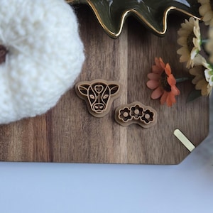 May include: Two brown plastic cookie cutters on a wooden surface. One cutter is shaped like a cow's head and the other is shaped like three flowers.