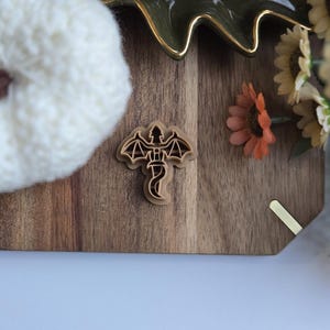 May include: A wooden cookie cutter shaped like a dragon with its wings outstretched. The cutter is on a wooden surface with a white knitted pumpkin and a bunch of flowers in the background.