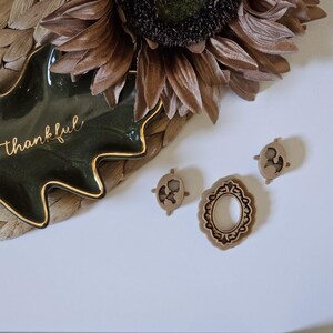 May include: A green ceramic leaf-shaped dish with a gold rim and the word "thankful" written in gold lettering. The dish is sitting on a woven straw mat. Three small wooden decorative pieces are on a white surface.