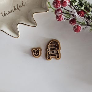 May include: Two wooden cookie cutters, one shaped like a car wearing a Santa hat and the other shaped like a bear. The cookie cutters are on a white surface.