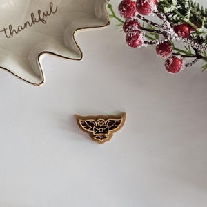 May include: A gold-coloured owl pin with a brown envelope in its talons. The owl is flying with its wings spread out. The pin is on a white surface.