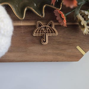 May include: A wooden cookie cutter shaped like an umbrella with a face. The cookie cutter is on a wooden surface.