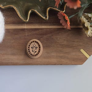 May include: A wooden oval-shaped ornament with a carved floral design. The ornament is on a wooden surface.