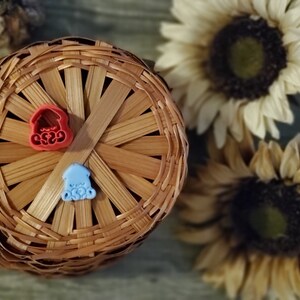May include: Two small plastic figurines, one red and one blue, are sitting on a woven basket. The figurines are shaped like houses with rounded roofs.