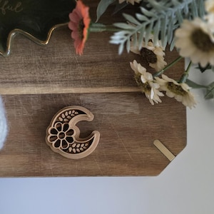 May include: A wooden crescent moon-shaped craft element with a floral design in the centre. The moon is light brown and sits on a wooden surface. Decorative flowers and a ball of white yarn are in the background.
