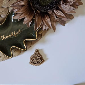 May include: A dark green leaf-shaped ceramic dish with gold trim and the word "thankful" in gold script. A small wooden pumpkin-shaped cookie cutter sits nearby. A dried sunflower and woven placemat are also visible.