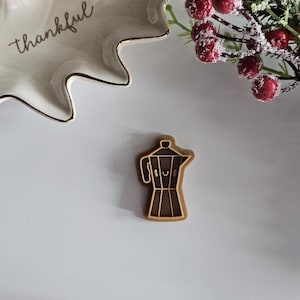 May include: A brown wooden cookie cutter shaped like a coffee pot with a smiling face. The cookie cutter is on a white surface.