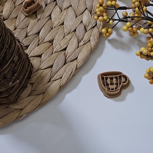 May include: A brown cookie cutter shaped like a coffee cup with a handle. The cookie cutter is on a white surface with a woven mat and yellow berries in the background.