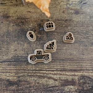 May include: Five cookie cutters on a wooden surface. The cutters are shaped like a Christmas tree, a pickup truck, a stack of presents, a single acorn, and a stack of three ornaments.
