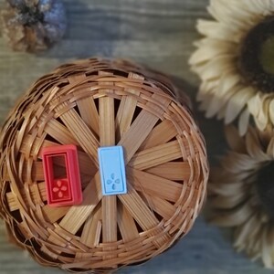 May include: Two small, decorative tiles with a floral design. The tile on the left is red with a white flower, and the tile on the right is blue with a white flower. The tiles are sitting on a woven basket.