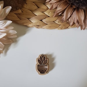 May include: A brown wooden cookie cutter shaped like a potted plant with a single stem and leaves. The cookie cutter is on a white surface.