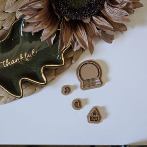 May include: A set of four brown cookie cutters in the shape of a snow globe, a crescent moon, a ghost, and a house. The cookie cutters are on a white surface.