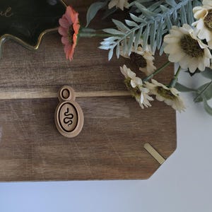 May include: A wooden pendant featuring a snake design rests on a wooden chopping board. The pendant is light brown with a dark brown snake illustration. The chopping board is surrounded by flowers and greenery.