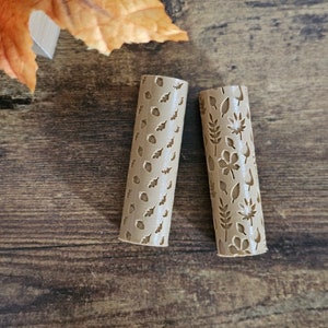 May include: Two beige cylindrical rolling pins with leaf and floral cutouts. The pins are made of a light brown material and are designed with various leaf and flower patterns. The background includes autumn leaves on a wooden surface.