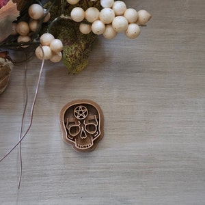 May include: A wooden skull cookie cutter with a pentagram design. The cutter is brown and has a detailed design.