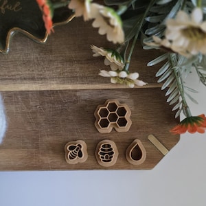 May include: Wooden bee-themed cookie cutters on a wooden cutting board. The set includes a honeycomb, bee, beehive, and teardrop shape. The board is decorated with flowers and greenery, with a white ball of yarn in the corner.