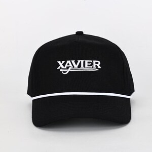 May include: Black baseball cap with the word "XAVIER" in white, arched above a white line and a stylized sword. A white rope detail trims the brim. The cap is a classic, casual accessory.