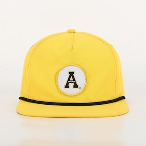 May include: A yellow flat-brimmed cap with a black rope detail along the brim. The cap features a circular patch with a black letter "A" on a white background, and the word "mountaineers" is written below the letter.