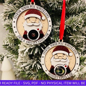 以下が含まれることがあります： 白地に赤いグリッターが施された丸い木製のオーナメントが2つ。それぞれのオーナメントには、カメラを持ったサンタクロースの漫画と、「北極圏監視本部」と「ジャックとアメリアのサンタカメラ」または「オリバーのサンタカメラ」という文字が書かれています。