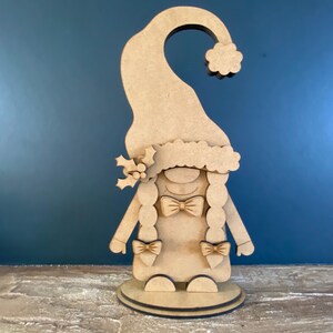 May include: A wooden gnome figurine with a red hat, a white fur trim, and a bow tie. The gnome is standing on a round base.