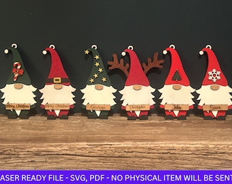 Puede incluir: Seis adornos de gnomo de Navidad de madera con diferentes diseños. Los gnomos llevan gorros rojos y verdes y tienen barbas blancas. Los adornos tienen nombres escritos en ellos: Feliz Navidad, Feliz Navidad, George, Christopher, John, Sam.