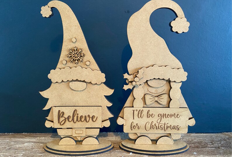 May include: Two wooden gnome figurines, one holding a sign that says "Believe" and the other holding a sign that says "I'll be gnome for Christmas".